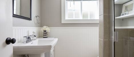 A bathroom with a white sink, a mirror, a window, and a shower area.