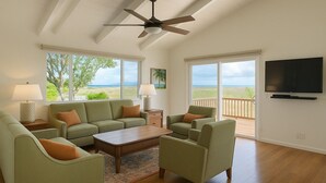 Ein geräumiges Wohnzimmer mit großem Fenster mit Blick auf einen Strand, einem Deckenventilator und einem an der Wand montierten Flachbildfernseher.