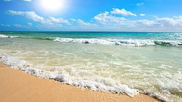 Uma praia com águas azul-claras, ondas e uma faixa de areia dourada sob um sol radiante.