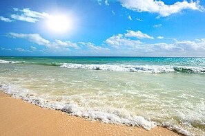 En strand med kristallklart blått vatten, vågor och en sandstrand under en strålande sol.