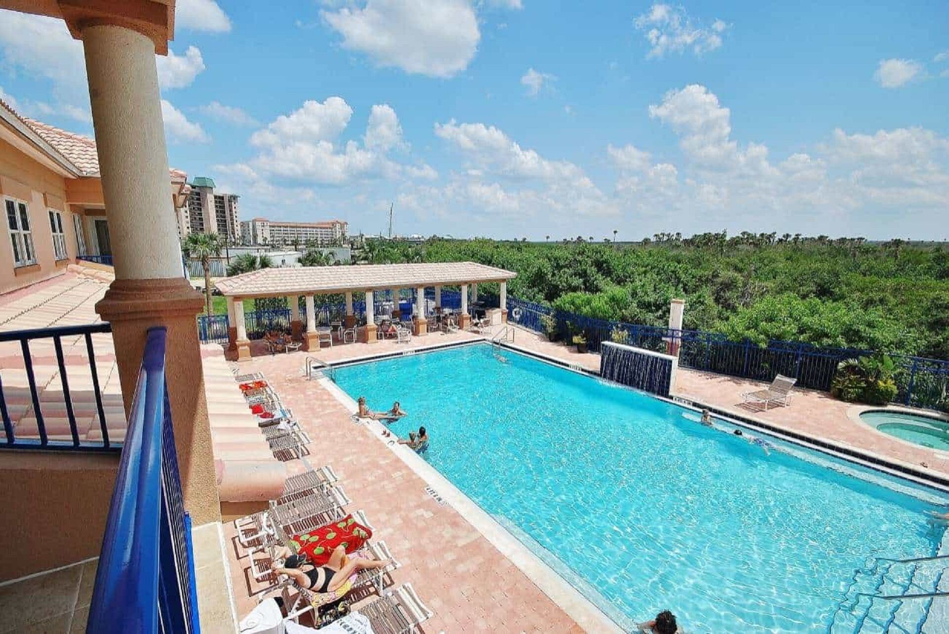 A swimming pool with lounge chairs and a covered seating area, surrounded by greenery and buildings.