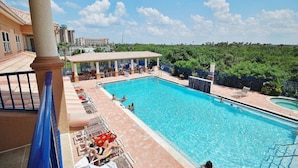 Piscina com espreguiçadeiras e área de estar coberta, cercada por vegetação e edifícios.