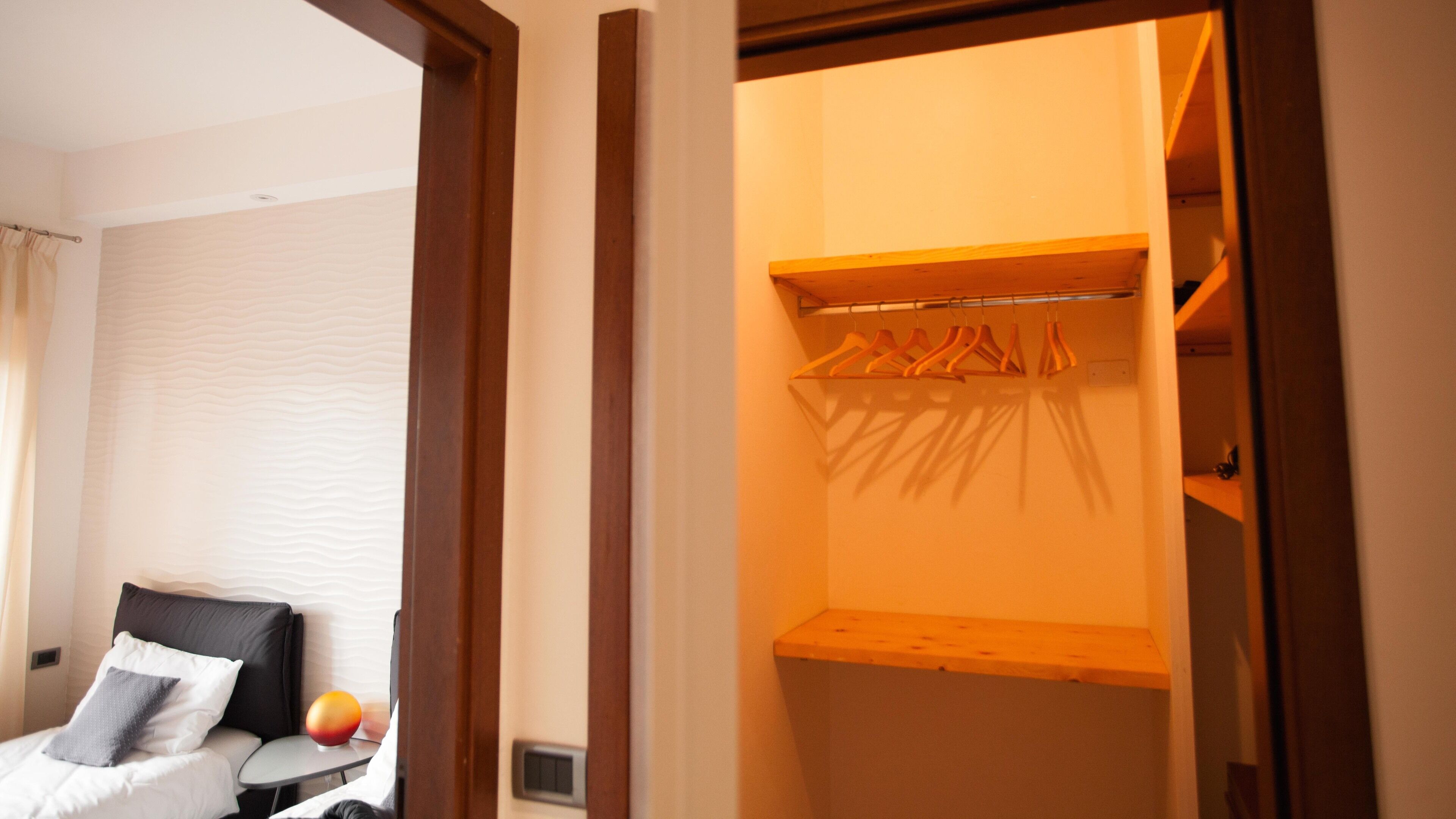 A hotel room with a bed, a bedside table, and a wardrobe with hanging clothes.