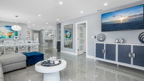 A modern living room with a dining area, a large painting of a sunset, and a sleek cabinet with decorative items. - Sea Breeze Paradise - Walk to Vanderbilt Beach and Mercato, Heated Pool Home (Naples)