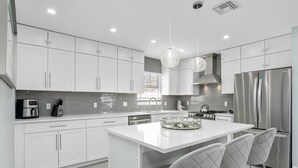 A modern kitchen with white cabinets, a stainless steel refrigerator, and a central island with bar stools. - Sea Breeze Paradise - Walk to Vanderbilt Beach and Mercato, Heated Pool Home (Naples)