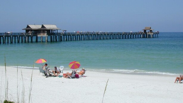 A beach with a pier extending into the water, featuring several beach umbrellas and people relaxing on the sand. - Sea Breeze Paradise - Walk to Vanderbilt Beach and Mercato, Heated Pool Home (Naples)
