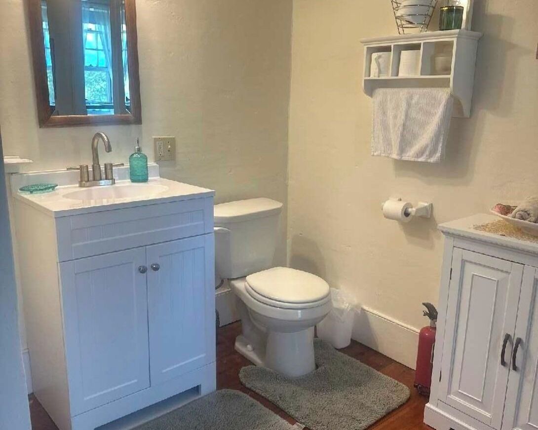 Un baño con lavamanos, inodoro y mueble blanco. Hay un espejo sobre el lavamanos y un perchero en la pared.