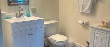 Un baño con lavamanos, inodoro y mueble blanco. Hay un espejo sobre el lavamanos y un perchero en la pared.