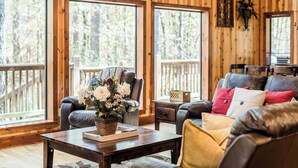 A cozy living room with a wooden interior, large windows, a leather sofa, and a coffee table with a floral arrangement. - Time Well Wasted- Spacious! Hot Tub,Shuffleboard,Playset,Free firewood! (Broken Bow)