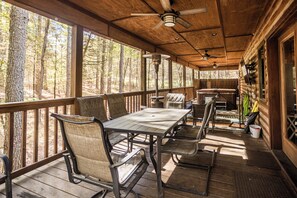 A covered outdoor area with a wooden deck, metal-framed tables and chairs, ceiling fans, and large windows offering a view of the surrounding forest. - Time Well Wasted- Spacious! Hot Tub,Shuffleboard,Playset,Free firewood! (Broken Bow)