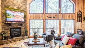 A cozy cabin interior with a stone fireplace, leather seating, and large windows offering a view of the forest. - Time Well Wasted- Spacious! Hot Tub,Shuffleboard,Playset,Free firewood! (Broken Bow)