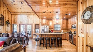 A wooden cabin interior with a dining area, kitchen, and bar stools. - Time Well Wasted- Spacious! Hot Tub,Shuffleboard,Playset,Free firewood! (Broken Bow)