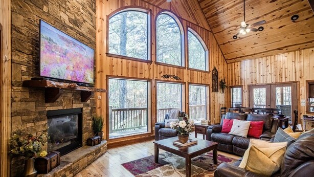A spacious living room with a stone fireplace, leather sofas, and large windows offering a view of the forest. - Time Well Wasted- Spacious! Hot Tub,Shuffleboard,Playset,Free firewood! (Broken Bow)