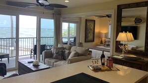 Ein Wohnzimmer mit einer Couch, einem Couchtisch und einer Küchenzeile mit Herd. Ein großes Fenster bietet Blick aufs Meer.