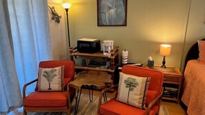 A hotel room with two red armchairs, a small wooden table, a lamp, a framed picture on the wall, and a bed with a pink blanket.