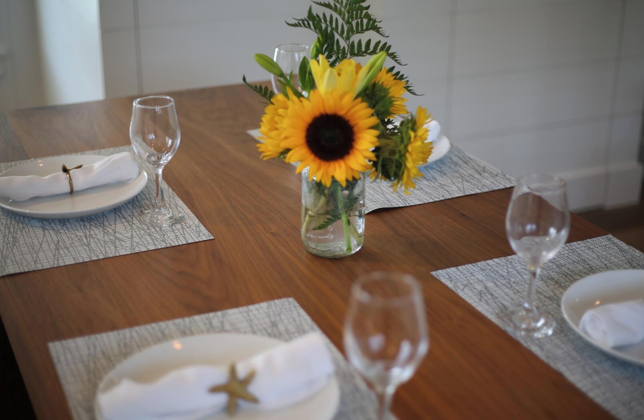 Una mesa de comedor dispuesta con platos blancos, servilletas y copas de vino, junto con un jarrón de girasoles y follaje verde.