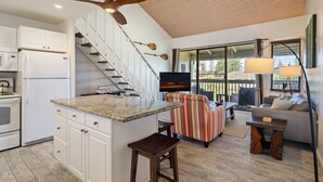 Eine moderne Küche mit weißen Schränken, einer Granit-Arbeitsplatte und einem eingebauten Backofen. Ein Hängelampenventilator aus Holz, eine Treppe mit weißen Geländern und ein großes Fenster mit Blick auf eine Terrasse sind ebenfalls vorhanden.