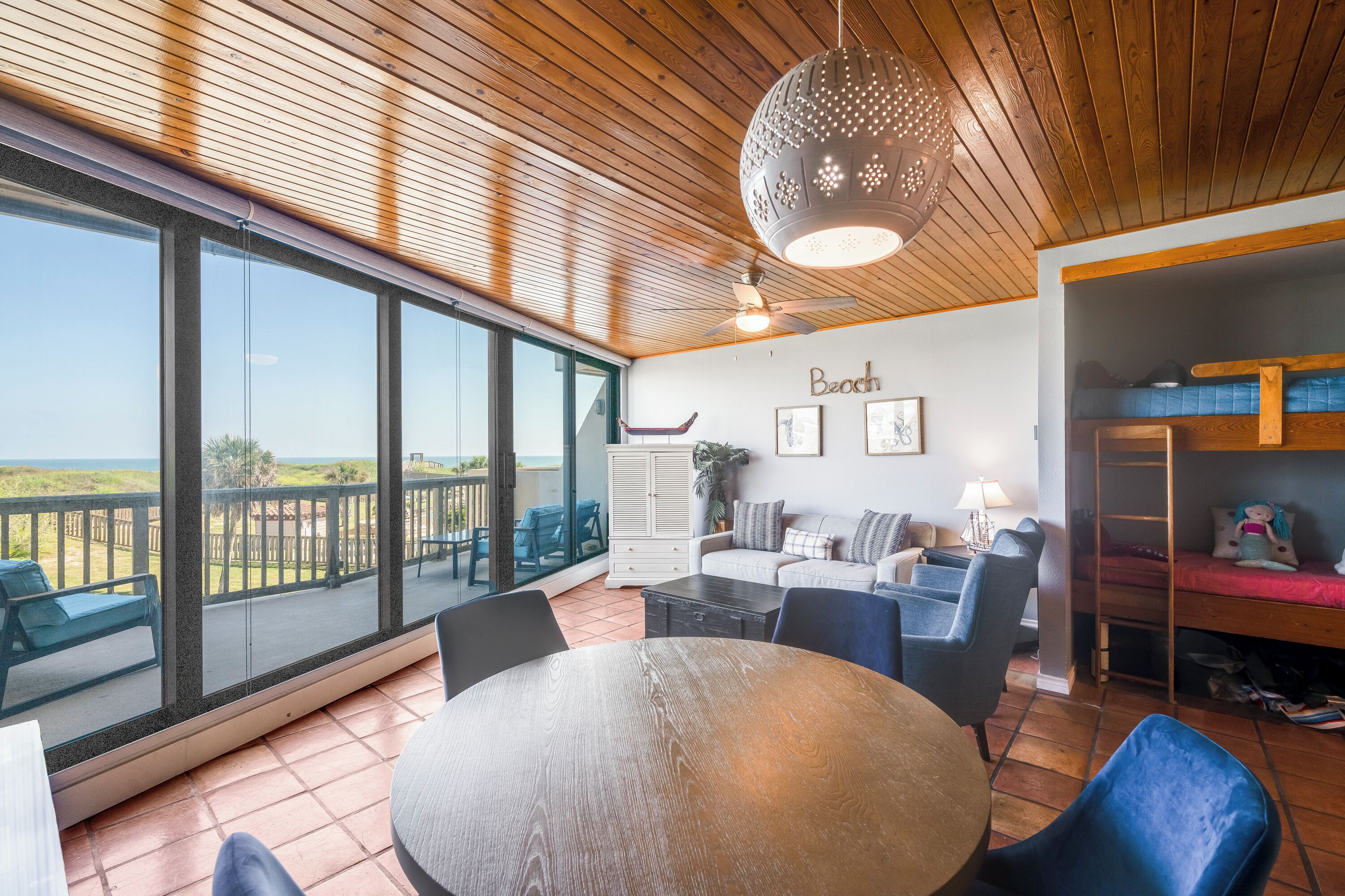 A spacious room with a large window overlooking a beach, a round wooden table, and a ceiling fan.