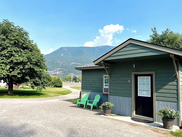 A small, single-story building with a dark green exterior, a front door, and a small porch area with two green Adirondack chairs.