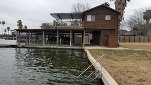 Una casa de madera con balcón que da a un cuerpo de agua, una terraza cubierta y un patio cercado.