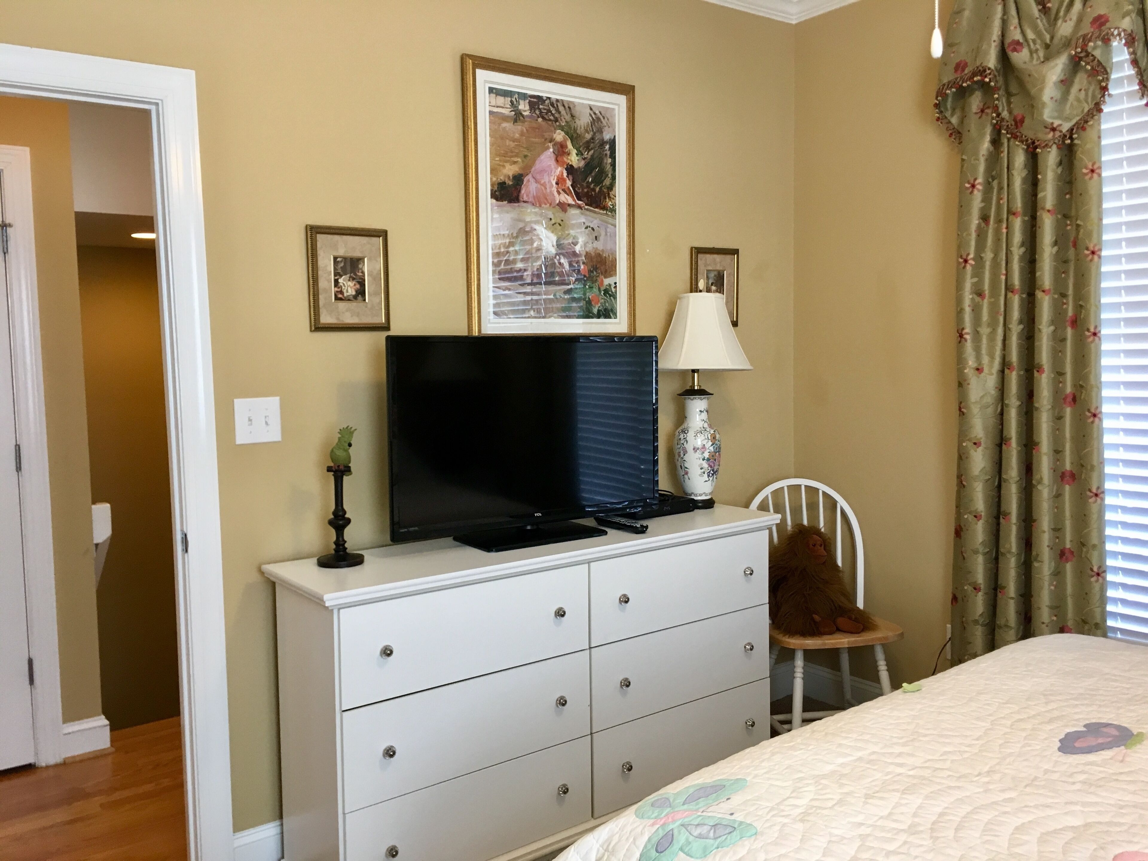 A bedroom with a television on a dresser, a lamp, a bed with a floral bedspread, and a window with curtains.