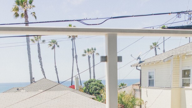 A balcony with two white chairs, overlooking a row of palm trees and a building with a sloped roof. - Beautiful & Clean!  White Water Views! (Oceanside)