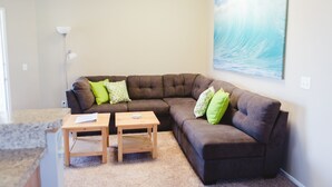 A living room with a brown sectional sofa, two wooden side tables, and a painting of a wave on the wall. - Beautiful & Clean!  White Water Views! (Oceanside)