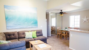 A living room with a large ocean wave painting, a brown sofa, a coffee table, and a dining area with wooden chairs and a glass-top table. - Beautiful & Clean!  White Water Views! (Oceanside)
