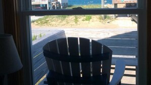 Vista desde una ventana, con una silla de madera en primer plano, una playa y edificios al fondo.