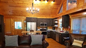 A cozy cabin interior with a wooden ceiling, a chandelier, a dining area, a kitchen with modern appliances, and leather furniture.
