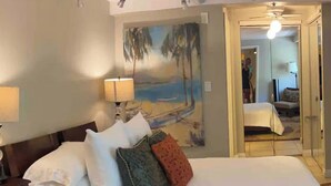 Un letto rifatto con lenzuola bianche e diversi cuscini, un comodino con una lampada e un quadro raffigurante una spiaggia.