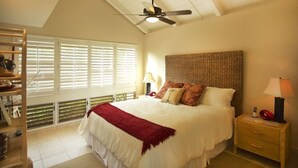 Une chambre avec un grand lit, un ventilateur de plafond, une table de chevet avec une lampe et une fenêtre avec des stores.