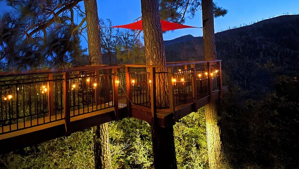 A wooden deck with string lights, surrounded by trees and a mountain backdrop. - PREMIUM TREE HOUSE  1.14-Acres  Hot Tub  Panoramic Lake Forest Mountain View (Julian)