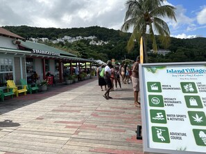 A wooden boardwalk with a Starbucks Coffee, green chairs, and a sign for Island Village activities. - Gordon's Beachfront Condo (Ocho Rios)