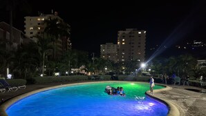 A nighttime swimming pool with a blue hue, surrounded by palm trees and buildings. - Gordon's Beachfront Condo (Ocho Rios)