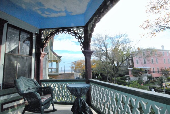 A balcony with a wicker chair and a small table, overlooking a street with buildings and trees.