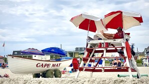 Une tour de surveillance avec des parasols rouges et blancs à rayures, une chaise de plage et un bateau nommé « GAPE MAY » sur le sable.