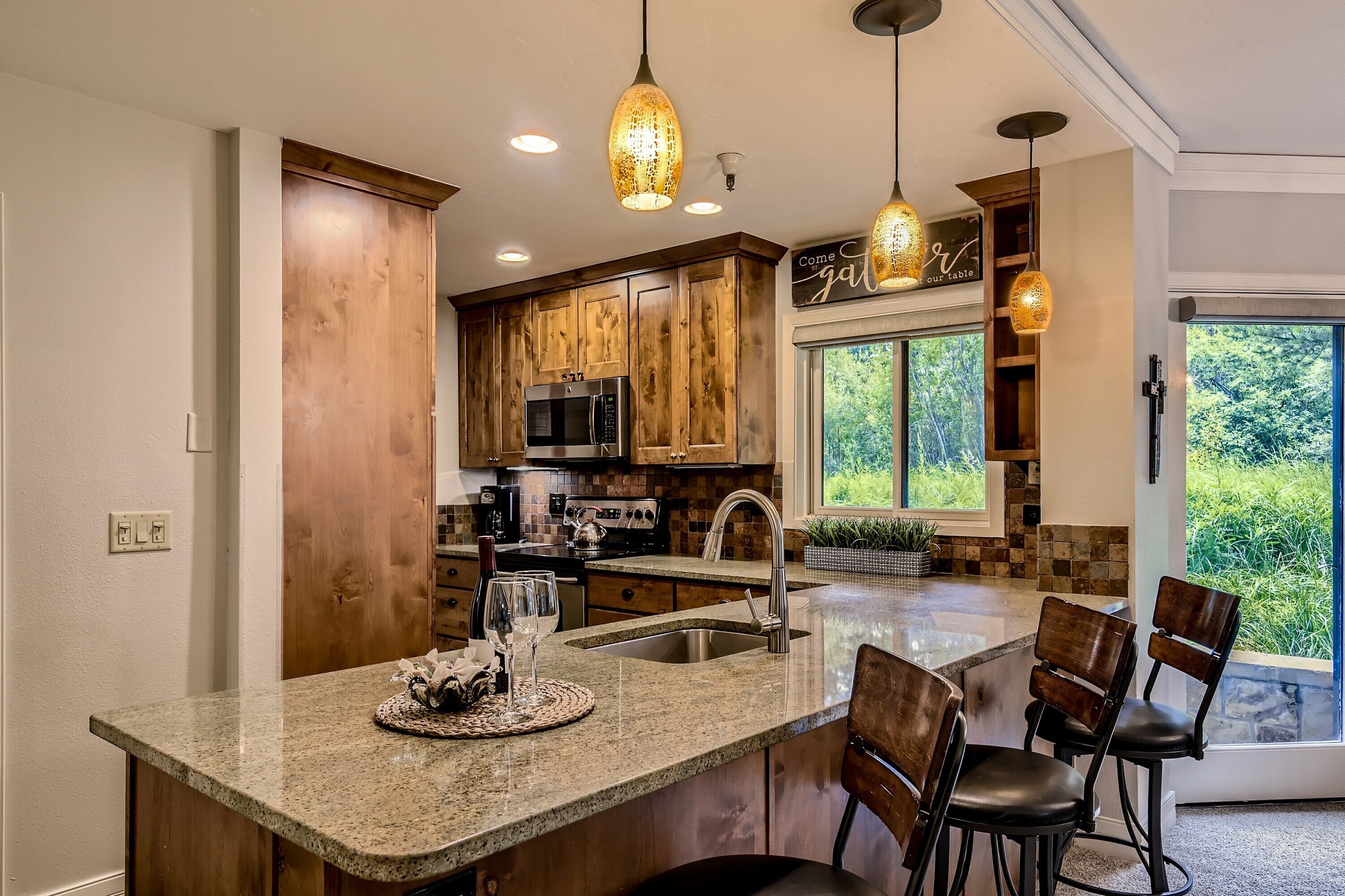 Una cocina con gabinetes de madera, un lavamanos de acero inoxidable y una isla grande con taburetes de bar. Hay lámparas colgantes sobre la isla y una ventana con vistas a la vegetación.