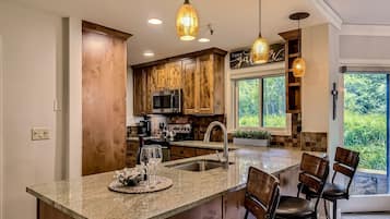 Una cocina con gabinetes de madera, un lavamanos de acero inoxidable y una isla grande con taburetes de bar. Hay lámparas colgantes sobre la isla y una ventana con vistas a la vegetación.