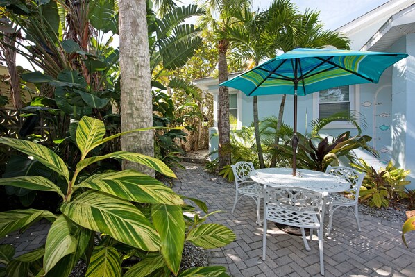 A patio with a white table and chairs, surrounded by lush tropical plants and a blue umbrella. - THE COTTAGE - 32 Steps to the Beach - Sleeps 2, drive to your door, Wifi, Grill. (Clearwater)