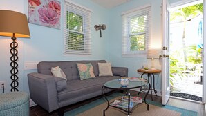 A living room with a grey sofa, a glass coffee table, a lamp, and a door leading to a garden. - THE COTTAGE - 32 Steps to the Beach - Sleeps 2, drive to your door, Wifi, Grill. (Clearwater)