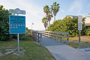 A wooden boardwalk leading to a beach with palm trees and a lifeguard sign warning of no lifeguard on duty.