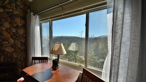 Ein Essbereich mit Steinwand, einem Tisch mit Stühlen, einer Lampe und Blick auf einen Wald durch große Fenster.