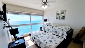 Quarto com uma janela grande, uma cama com colcha florida, uma escrivaninha com abajur e um ventilador de teto.