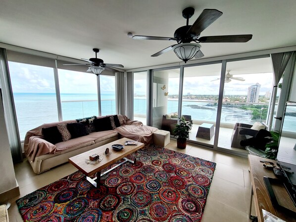 A spacious living room with a large window overlooking the sea, a ceiling fan, and a colorful rug.