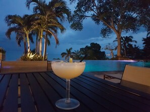 A cocktail on a wooden table with a pool and palm trees in the background.