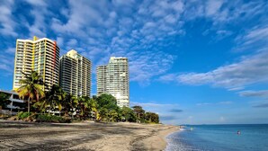Uma praia com edifícios altos, palmeiras e céu claro.