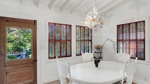 A dining area with a round table, white chairs, a chandelier, and windows with wooden shutters. - Authentic German Sunday House! Location, Outdoor Fireplace & Upscale Decor! (Fredericksburg)