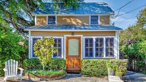 A two-story house with a front door, windows, and a stone pathway leading to a white chair. - Authentic German Sunday House! Location, Outdoor Fireplace & Upscale Decor! (Fredericksburg)