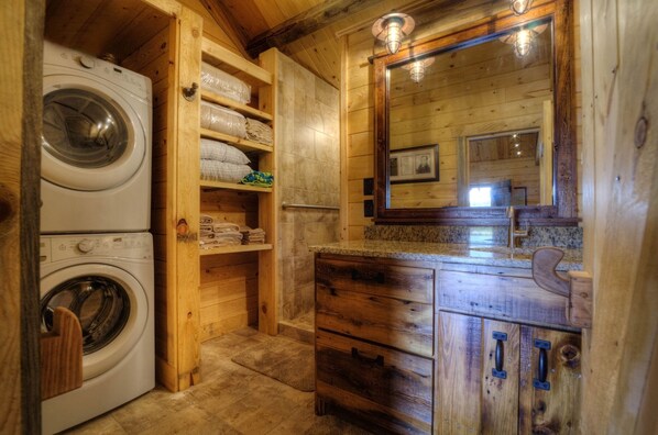 A wooden cabin interior with a washing machine, wooden shelves with folded towels, a granite countertop, and a mirror with a framed photo.